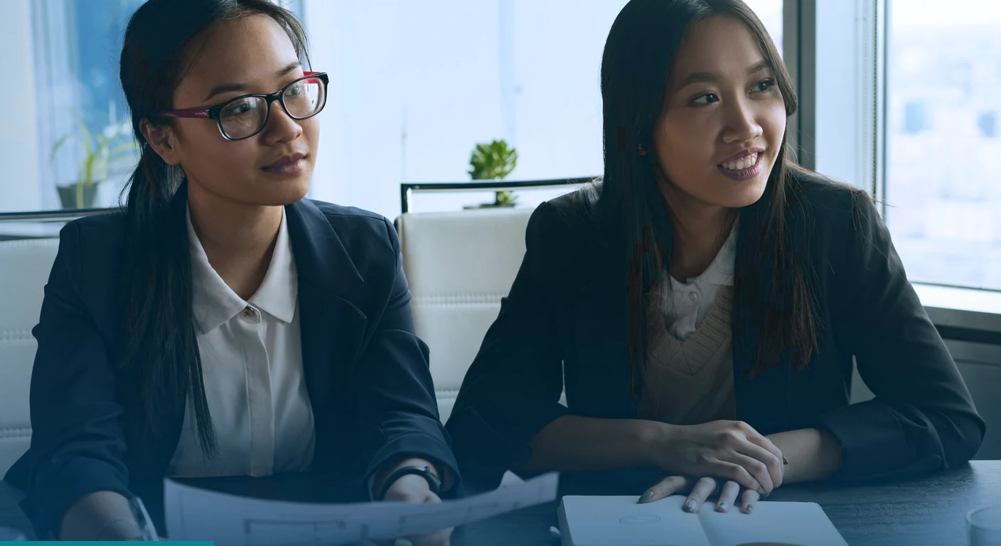Two colleagues in blazers at a meeting table, one holding printed design layouts