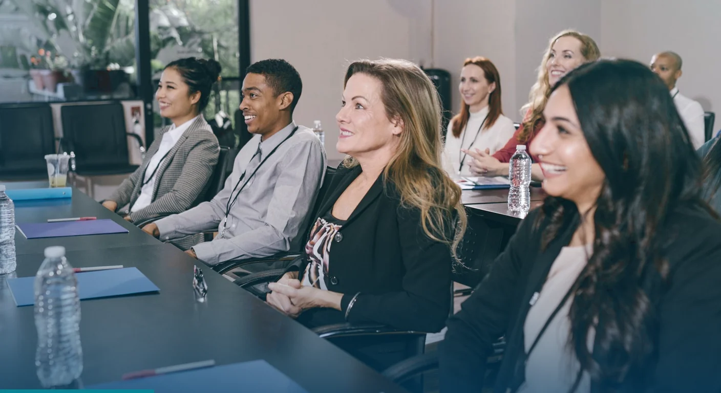 Smiling professionals with lanyards seated along a conference table, listening to a session
