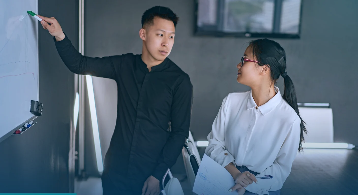 Man writing on a whiteboard while a colleague holding papers looks on