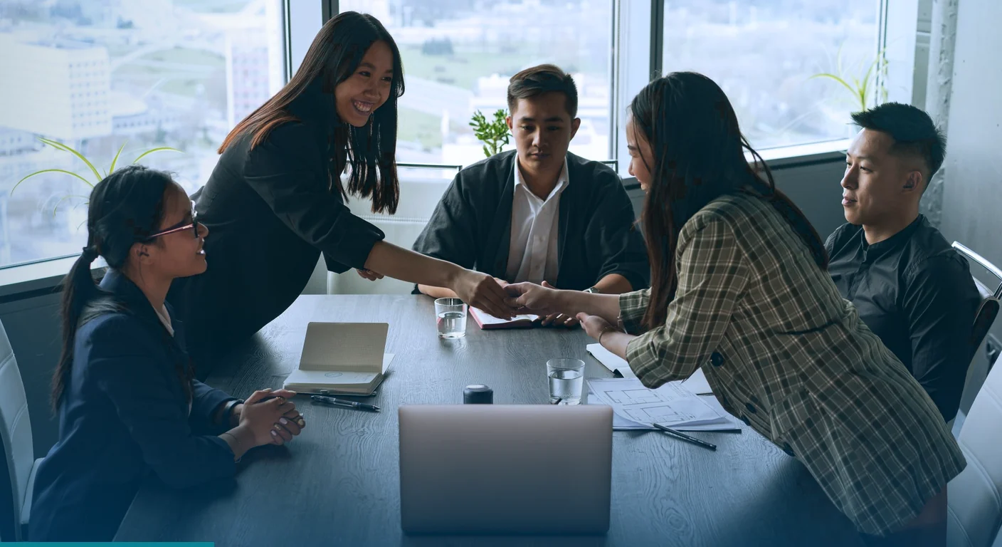 Team of colleagues shaking hands across a meeting table beside a bright office window