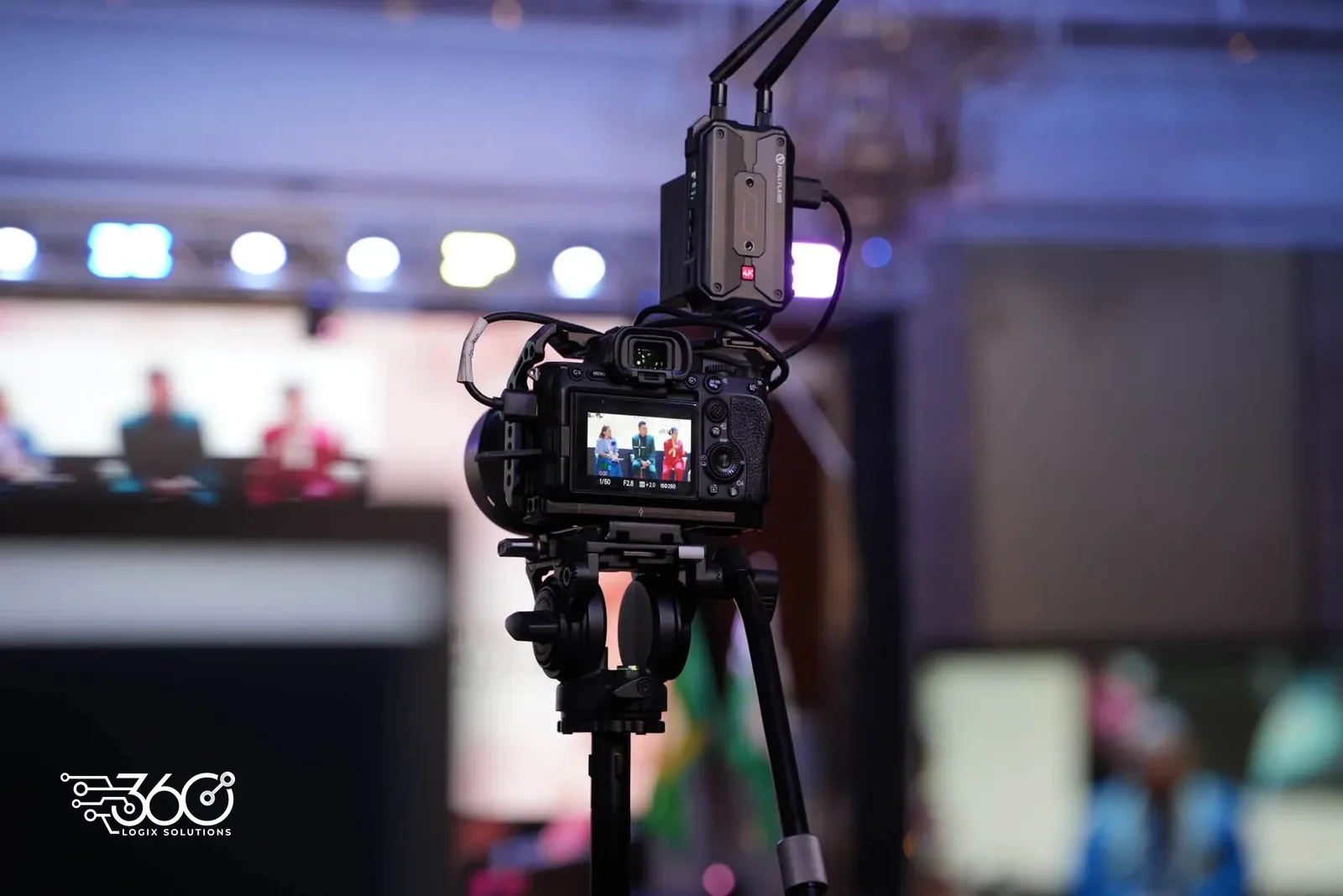 Production camera on a tripod at the PAPSHPI Aesthetic Summit 2026, its screen framing the speakers on stage, with stage lights glowing in the background.